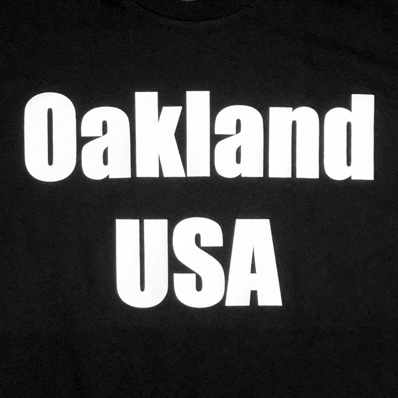 Close-up of large white Oakland USA wordmark logo on the chest of a black t-shirt.