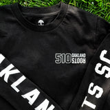 Close up of ¾ top view of black long sleeve t-shirt with Oakland Roots 510 logo on the chest, arms crossed over the chest on grass. 