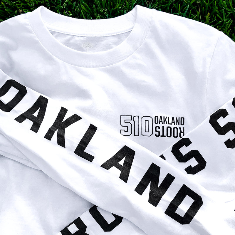 Close up of long sleeves with black printing saying Oakland in capital letters folding over the front of white t-shirt with 510 Oakland Roots logo.