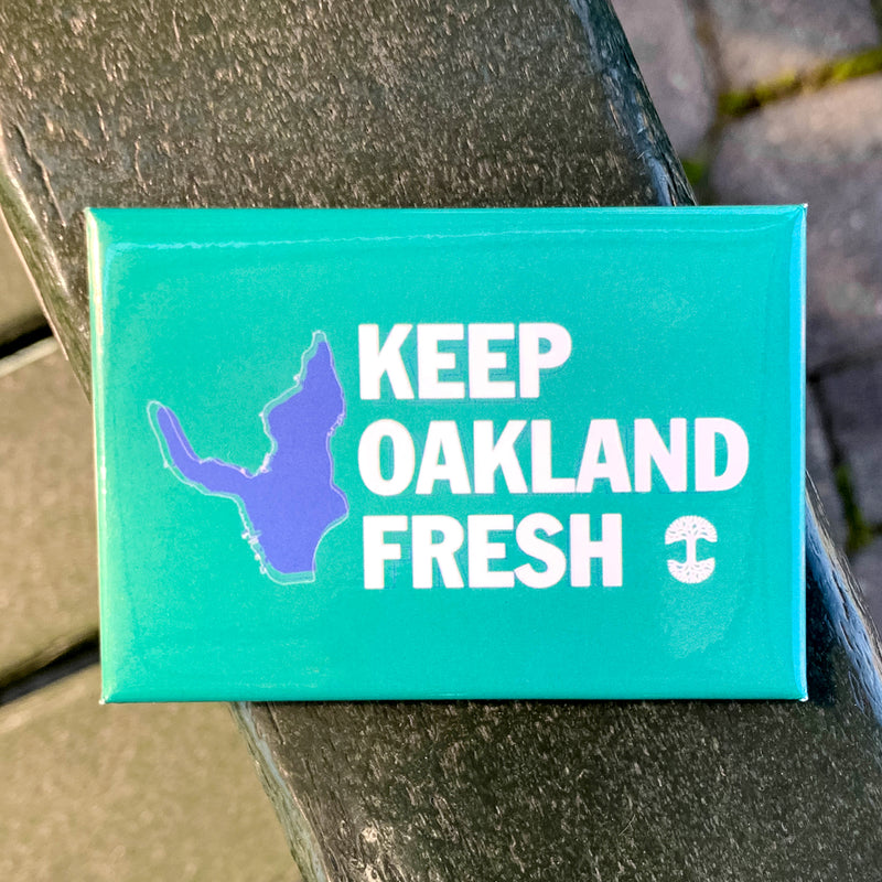 Green magnet with blue lake and KEEP OAKLAND FRESH wordmark and white Oaklandish tree logo outdoors.