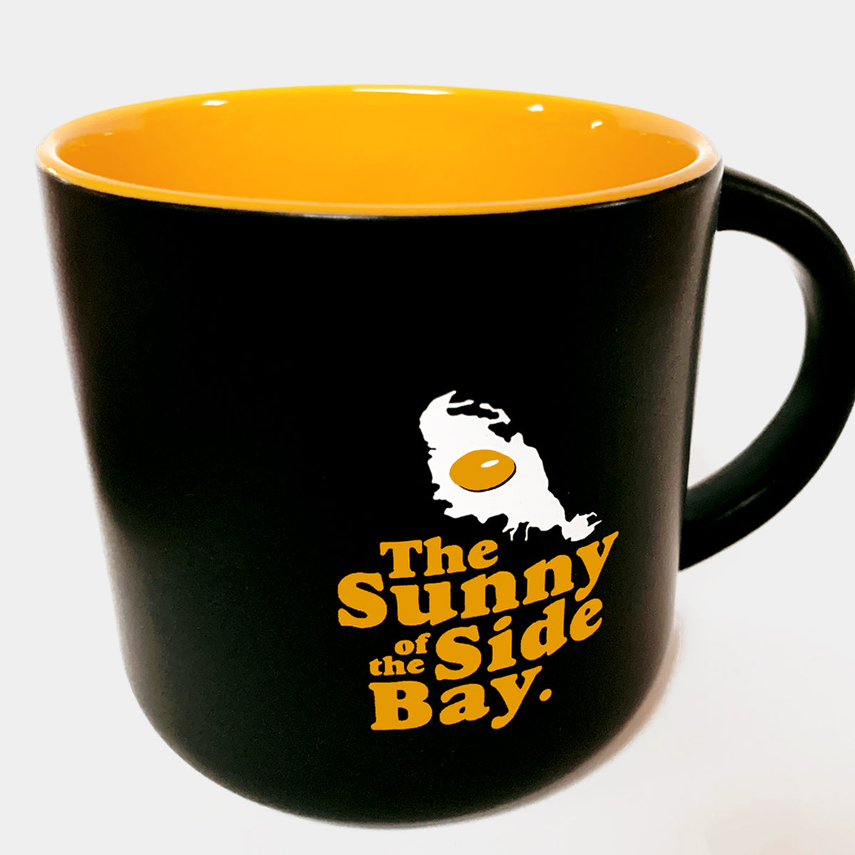 Black mug with yellow inside with picture of Oakland shaped sunny side up fried egg and words The Sunny Side Of The Bay.
