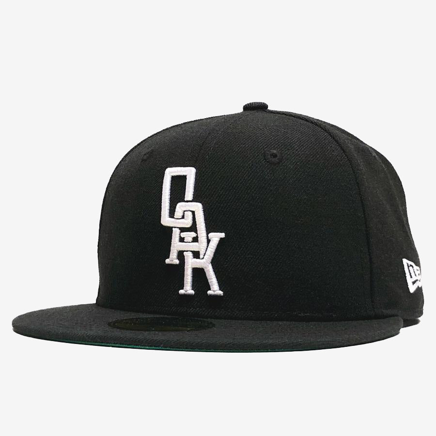 The New Era OAK 59FIFTY Fitted Cap by Oaklandish is a sleek, modern hat with a black fitted design and a flat brim, featuring the OAK logo embroidered in white stacked letters on the front. The side of the cap showcases the New Era logo in white.