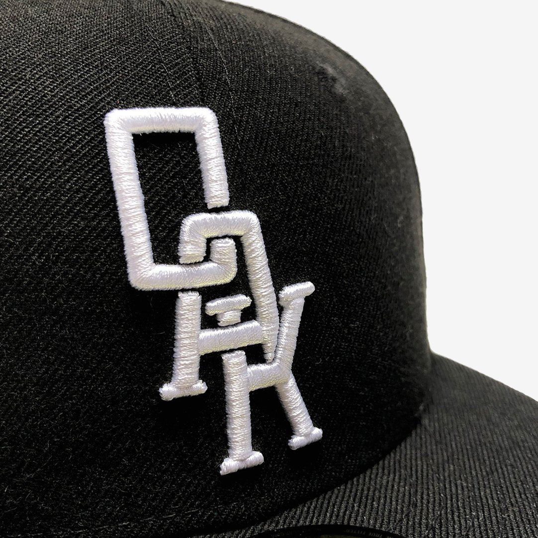 Close-up image of a black New Era OAK 59FIFTY Fitted Cap by Oaklandish, featuring a white interlocking "O", "A", and "K" logo embroidered on the front. The plain white background accentuates the cap's intricate details, showcasing perfectly overlapping vertically arranged letters.