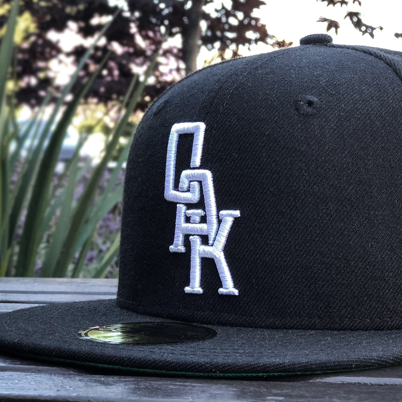 Close-up of a black New Era OAK 59FIFTY Fitted Cap by Oaklandish resting on a wooden surface outdoors. The cap features the intricate OAK logo in white, stacked vertically on the front panel. The background shows blurred greenery and trees, indicating a park or garden setting.