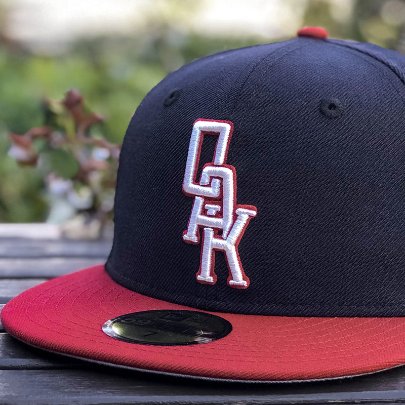 Close-up of a navy New Era cap with red visor and white embroidered OAK wordmark on the crown on a picnic table.