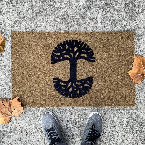 Brown fiber door mat with black Oaklandish tree logo on cement with fall leaves on a doorstep with two feet.