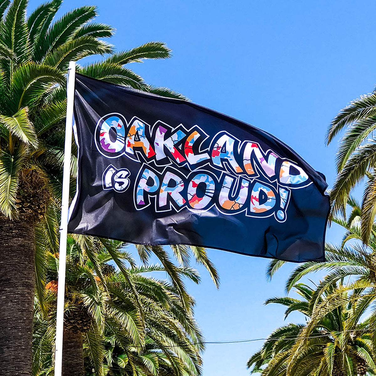 An Oaklandish "Oakland is Proud Flag" with bold, colorful text reading "OAKLAND IS PROUD!" waves in the wind. Set against tall palm trees and a clear blue sky, the letters are decorated with patterns and images like hearts and stripes. The vibrant theme echoes accessories seen in an Oakland mural by Del Phresh.