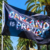 Close-up of white, red, blue, orange “OAKLAND IS PROUD!” wordmark on a black flag outdoors on a flag pole.