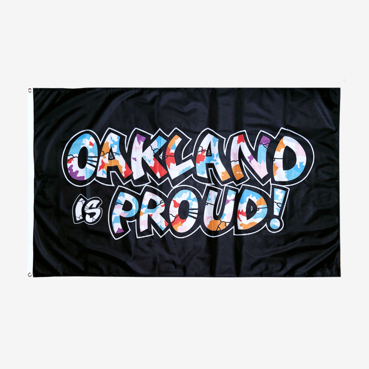 Introducing the Oaklandish "Oakland is Proud Flag" – a black flag with the words "OAKLAND is PROUD!" displayed in bold, colorful graffiti-style letters, evoking the essence of an iconic Oakland mural. The word "OAKLAND" bursts with vibrant colors and patterns, while "is PROUD!" features a primarily white aesthetic with color accents. Metal grommets on the left side facilitate easy hanging as an accessory.