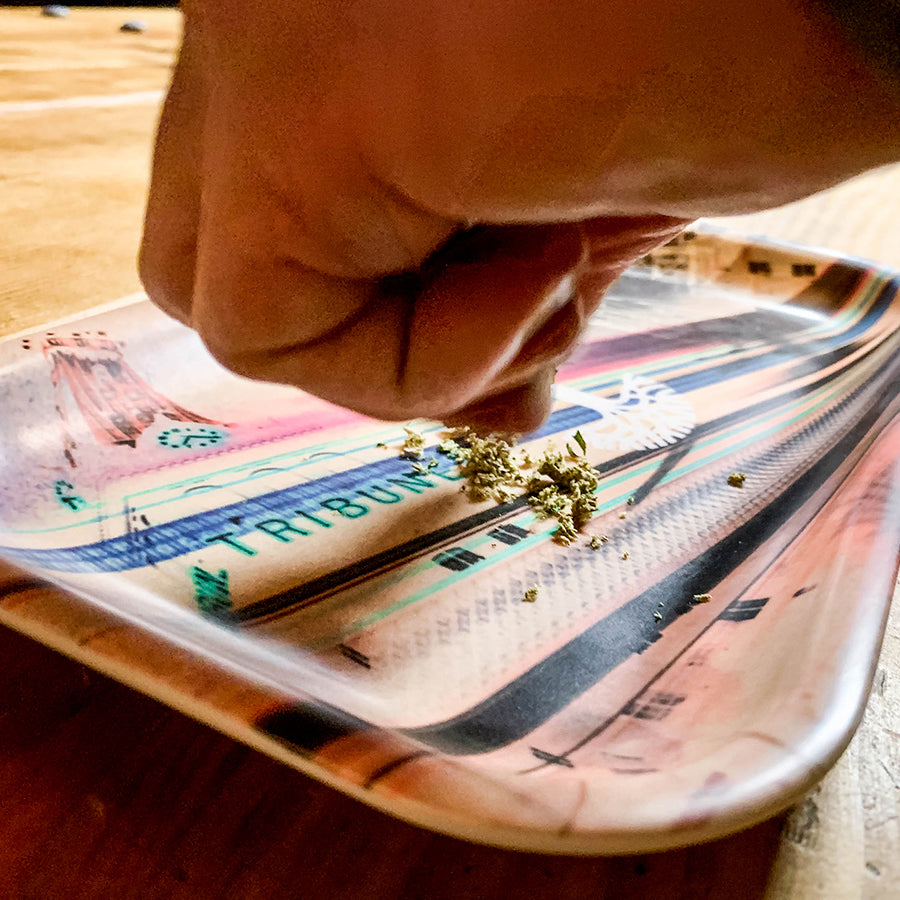 Close-up of a hand holding a small amount of ground material, possibly herbs or cannabis, over a colorful, rectangular multi-purpose tray with a vibrant pattern and the word "Neon High" by Oaklandish visible. The melamine composite tray rests on a wooden surface. The natural and soft lighting highlights the hand and the contents on the tray.
