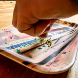 Hand adding marijuana to a rectangle melamine rolling tray with a black Oaklandish tree logo in the center.