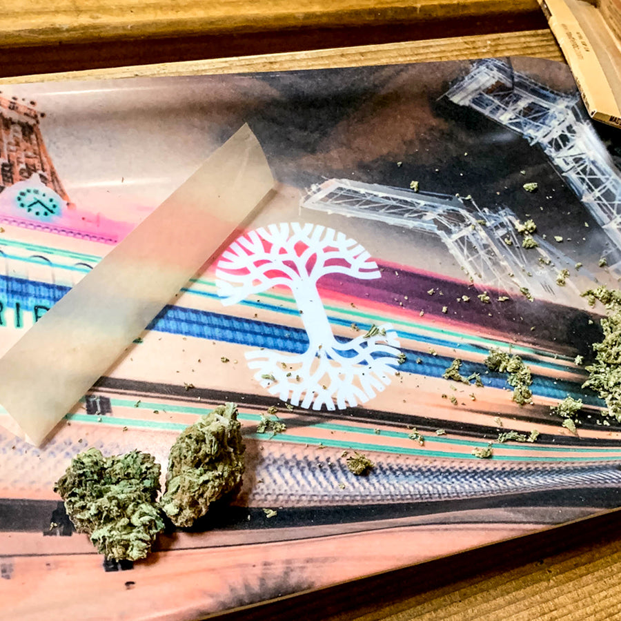 A rolling tray with marijuana buds, a half-rolled joint in partially translucent paper, and scattered marijuana fragments. The Neon High multi-purpose tray by Oaklandish features colorful artwork with a white tree design. The wooden surface underneath is also visible, making it a stylish addition to your accessories collection.