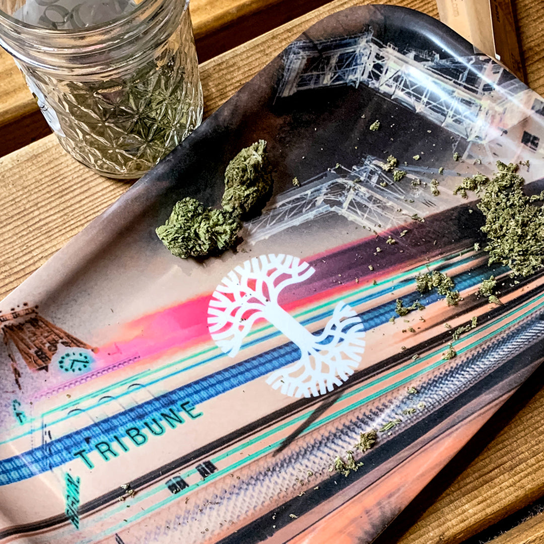 A rectangular, melamine composite rolling tray named "Neon High" by Oaklandish featuring abstract art and urban elements, including the word "TRIBUNE" in turquoise. The multi-purpose tray is perfect for holding cannabis buds and ground material. A glass jar containing cannabis is partially visible on a wooden surface nearby.