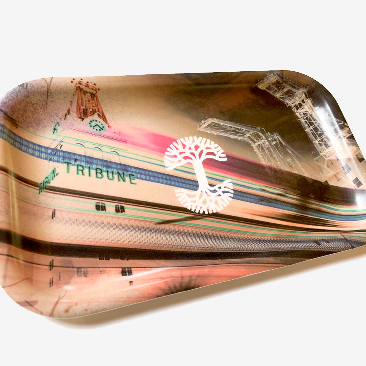 The "Neon High" by Oaklandish is a rectangular, multi-purpose tray showcasing a collage artwork. The design features abstract cityscape elements, a tree symbol at the center, and the word "TRIBUNE" on a building. Made from durable melamine composite, its backdrop combines warm and cool hues with blurred lines suggesting motion.