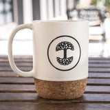 Cream colored rustic ceramic mug with black Oaklandish tree logo in a circle and cork bottom outdoors on a picnic table.