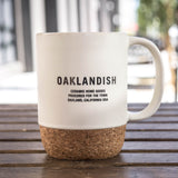 OAKLANDISH CERAMIC HOME GOODS PROCURED FOR THE TOWN on a cream colored ceramic mug with cork bottom outdoors on a picnic table.