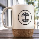 Cream colored rustic ceramic mug with black Oaklandish tree logo in a circle and cork bottom outdoors on a picnic table.