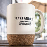 OAKLANDISH CERAMIC HOME GOODS PROCURED FOR THE TOWN on a cream colored ceramic mug with cork bottom outdoors on a picnic table.