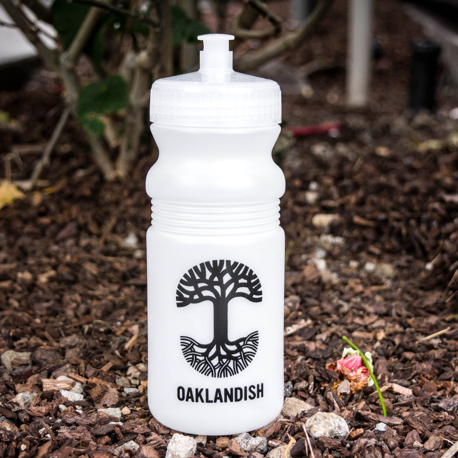 Frost white bicycle water bottle with a black Oaklandish tree logo and wordmark in a wooded outdoor area.