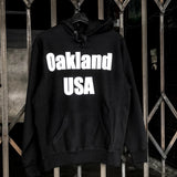Black hoodie with a large white Oakland USA wordmark logo on the chest hanging on an outdoor fence.