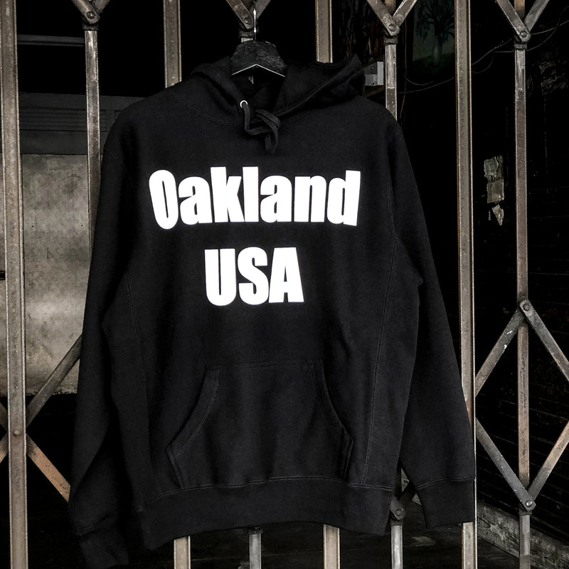 Black hoodie with a large white Oakland USA wordmark logo on the chest hanging on an outdoor fence.