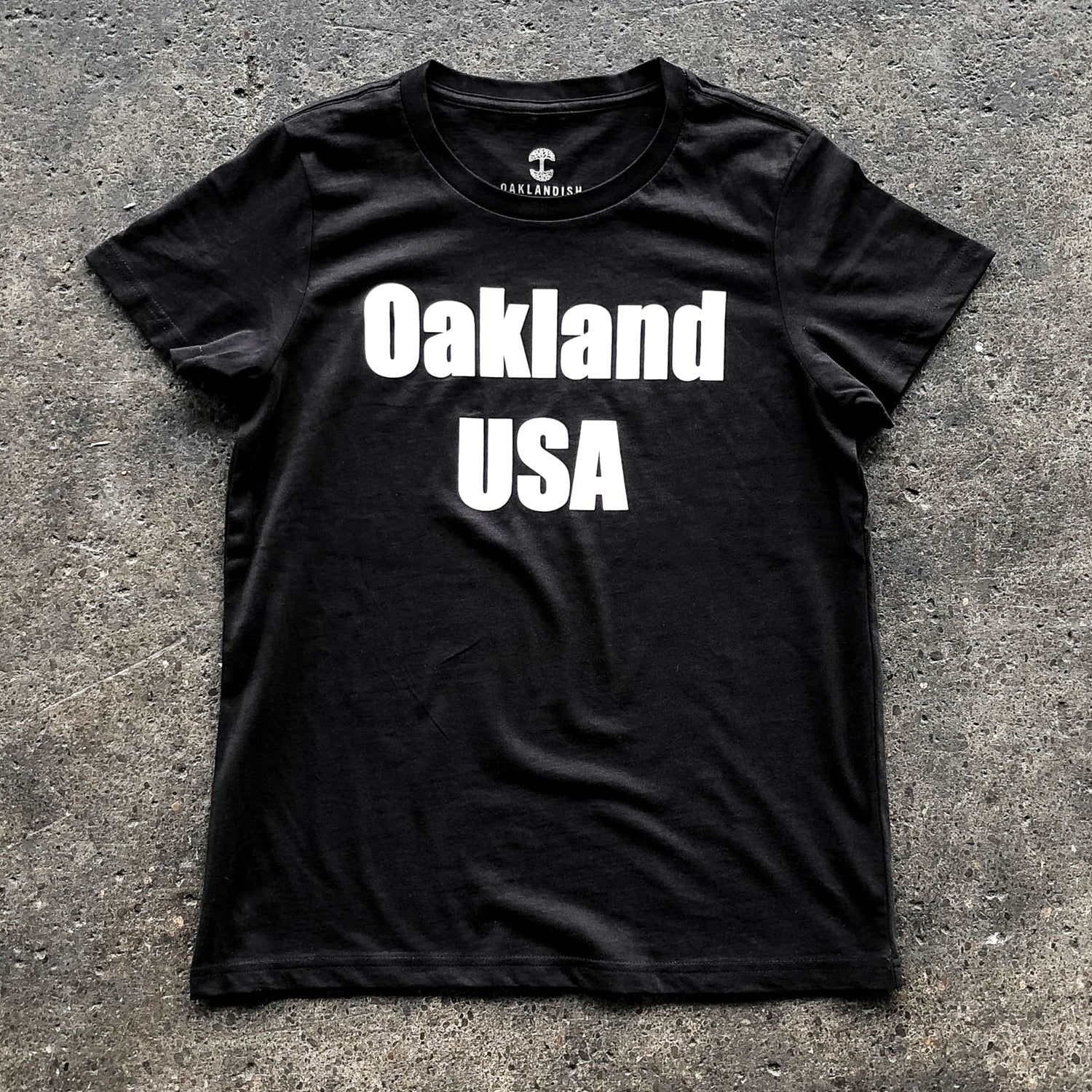 A women’s black t-shirt with a large white Oakland USA wordmark logo on the chest laying on asphalt.