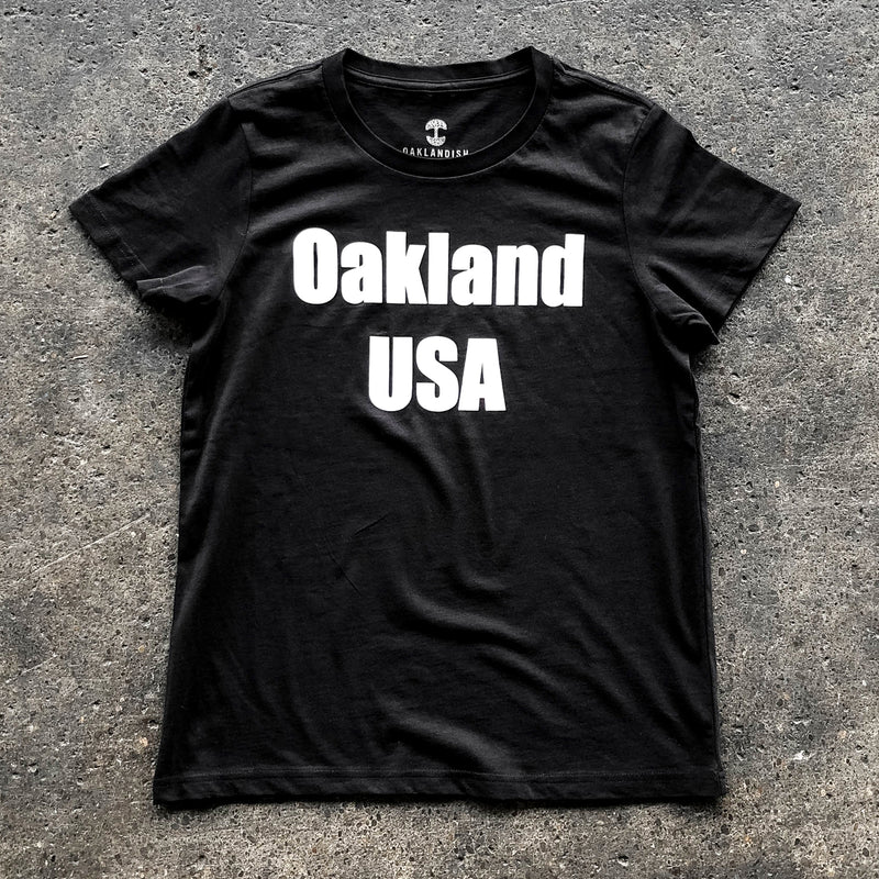 A women’s black t-shirt with a large white Oakland USA wordmark logo on the chest laying on asphalt.