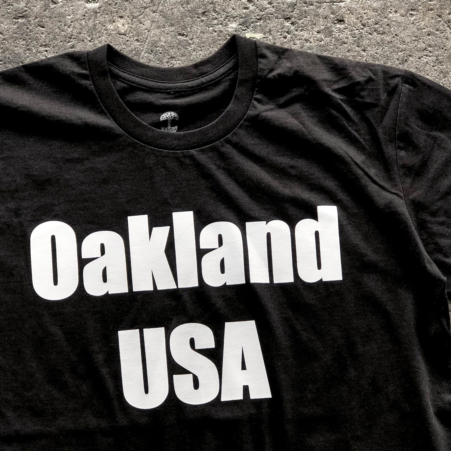 Close-up of large white Oakland USA wordmark logo on the chest of a black t-shirt laying on asphalt.