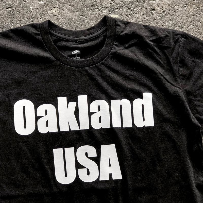 Close-up of large white Oakland USA wordmark logo on the chest of a black t-shirt laying on asphalt.