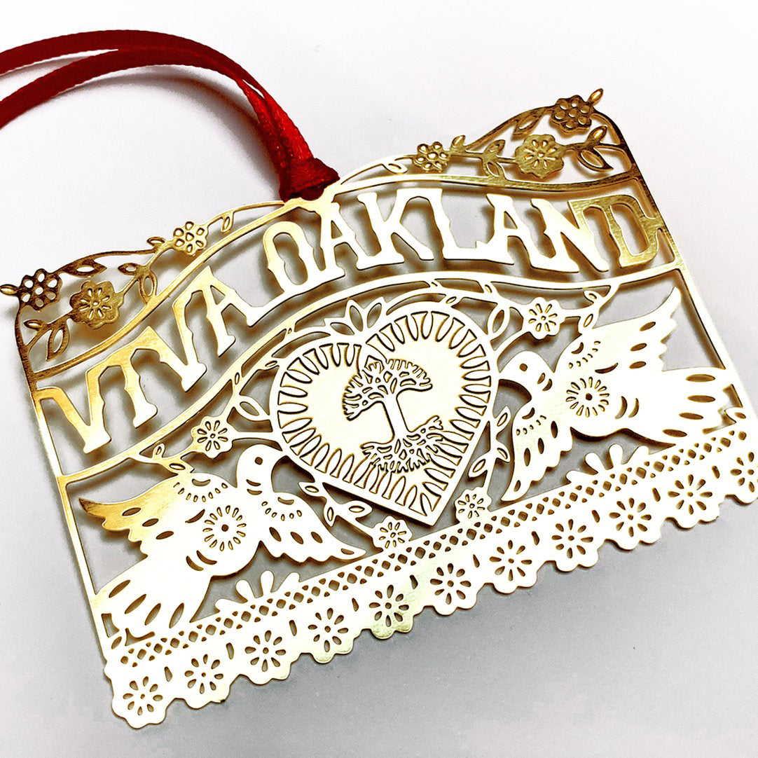 Close-up of brass ornament with Viva Oakland design with heart, birds and flowers and a red string.