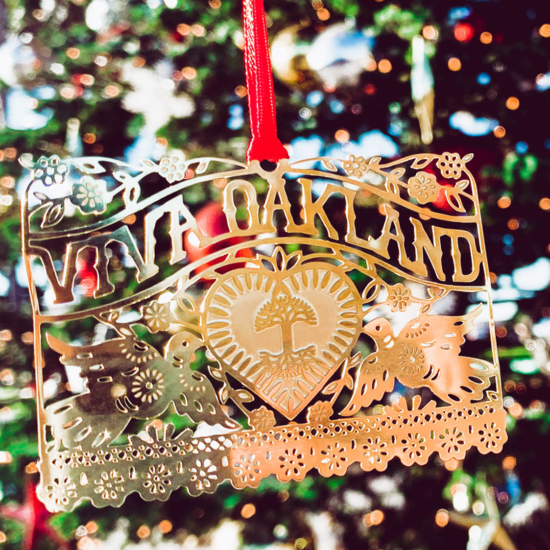 Front side of brass ornament with Viva Oakland design with heart, birds and flowers and a red string hanging on a Christmas tree.