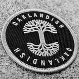 Round black circle patch with embroidered silver Oaklandish tree logo in the center and Oaklandish wordmarks above and below on ashphalt.
