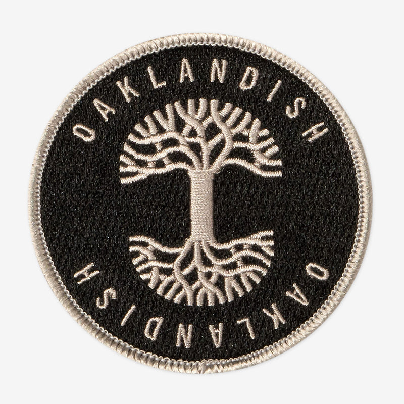 Round black circle patch with embroidered silver Oaklandish tree logo in the center and Oaklandish wordmarks above and below.