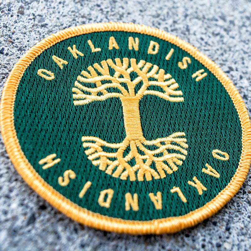 Round green circle patch with embroidered yellow Oaklandish tree logo in the center and Oaklandish wordmarks above and below on asphalt.