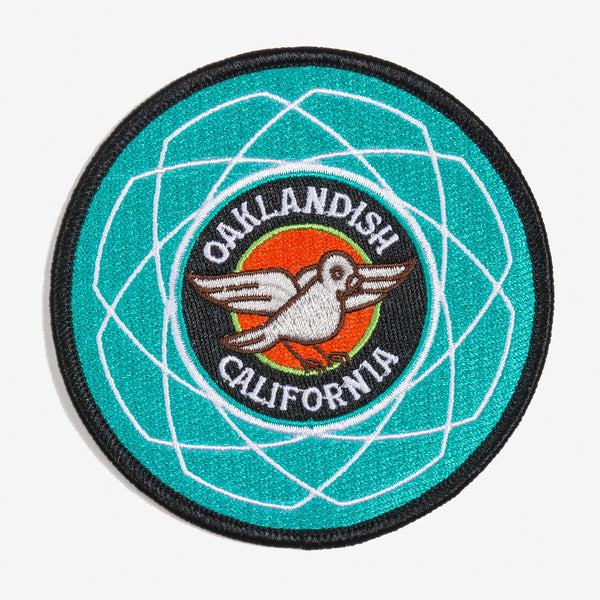 Round embroidered iron-on patch with turquoise blue outer rim, white pigeon in middle orange and grey circles, and “Oaklandish California” wordmark. 