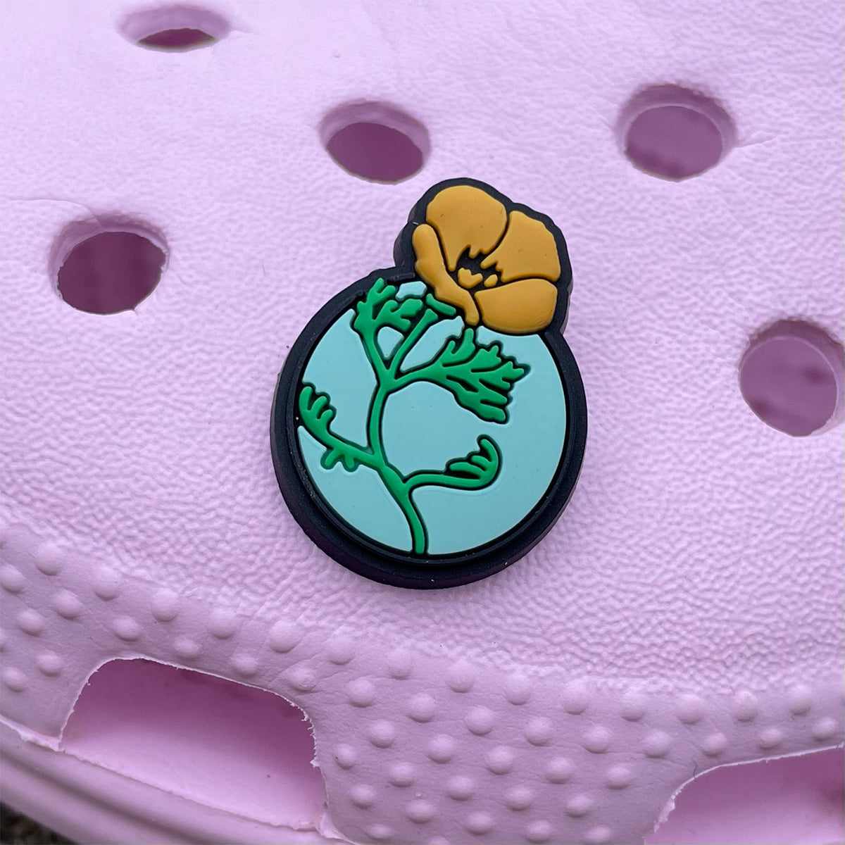 Close up of California poppy shoe charm with a yellow poppy, green stem, round blue background and a black base on a pink croc.