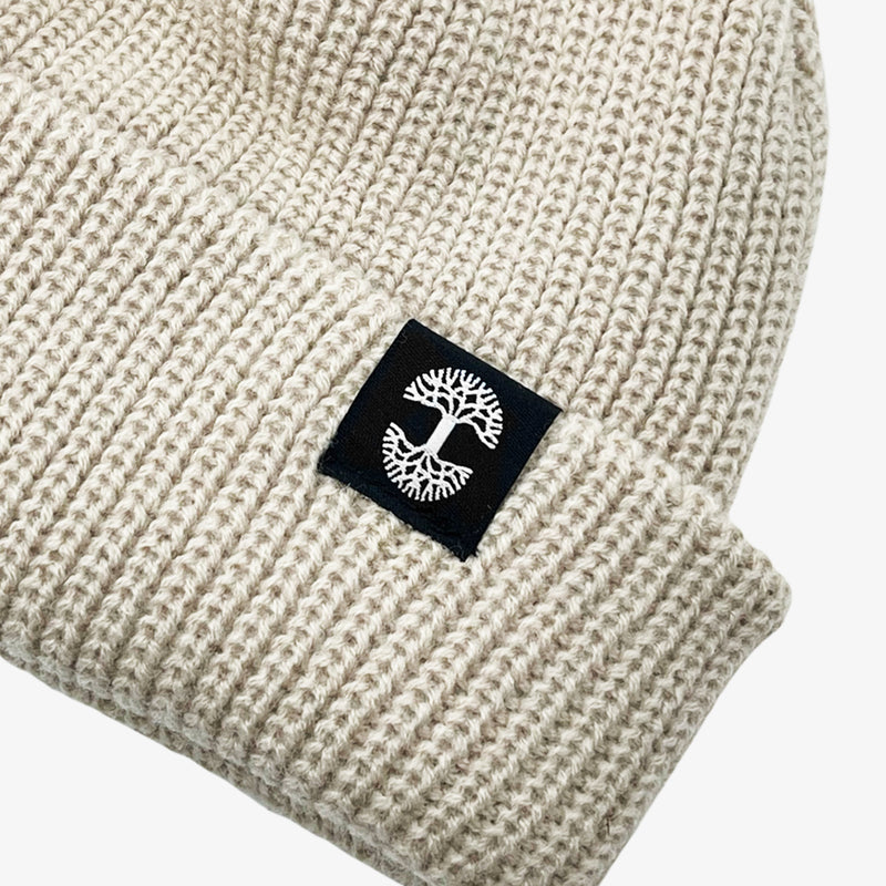 Close up of black and white Oaklandish tree logo tag on the left wear side of a shallow fit cream cuffed beanie.