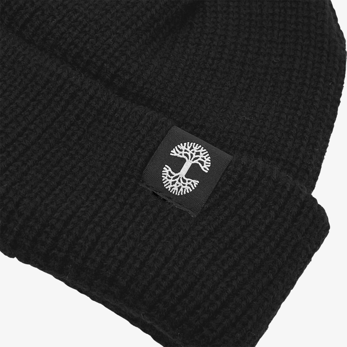 Close up of black and white Oaklandish tree logo tag on the left wear side of a shallow fit black cuffed beanie.