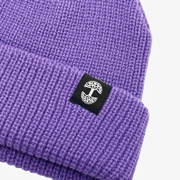 Close up of black and white Oaklandish tree logo tag on the left wear side of a shallow fit purple cuffed beanie.