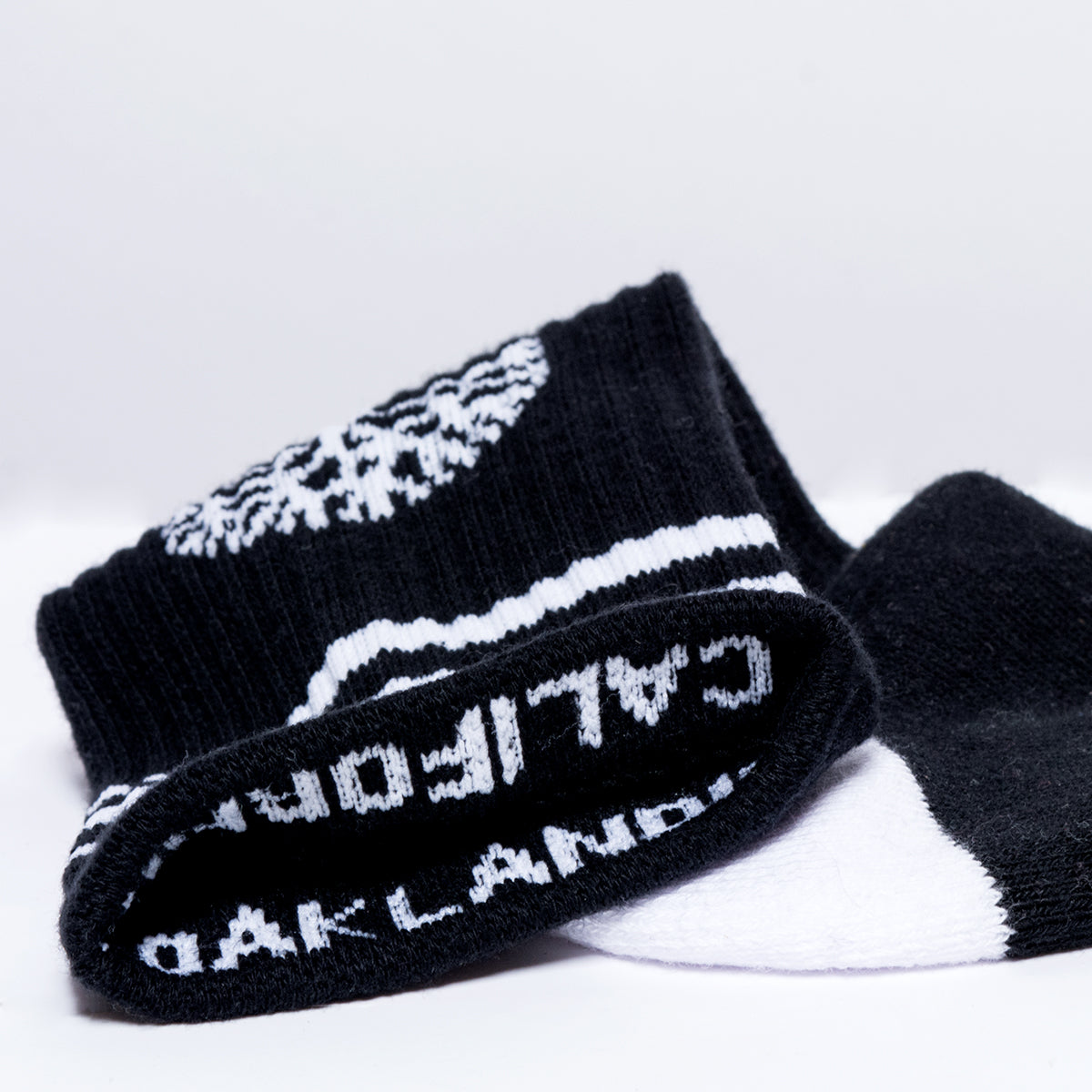 Top of a single black crew sock exposing OAKLANDISH and CALIFORNIA wordmarks on the inside cuff.