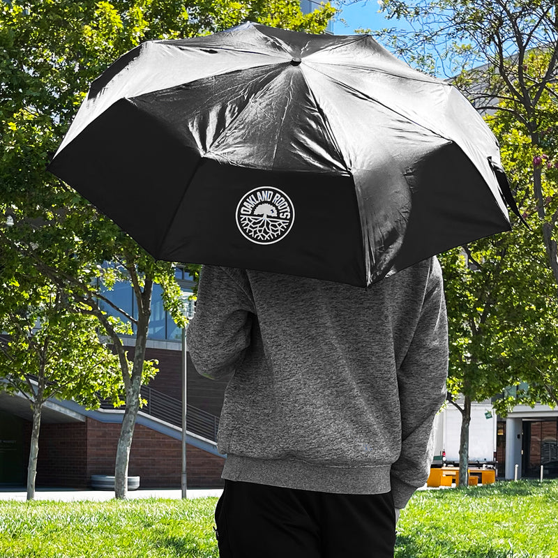 A man standing outside in a park on a sunny day; back to the camera carrying an open black umbrella with a round white Oakland Roots SC logo.