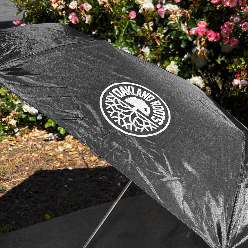 Open black umbrella outside with round with white Oakland Roots SC logo lying on the ground. 