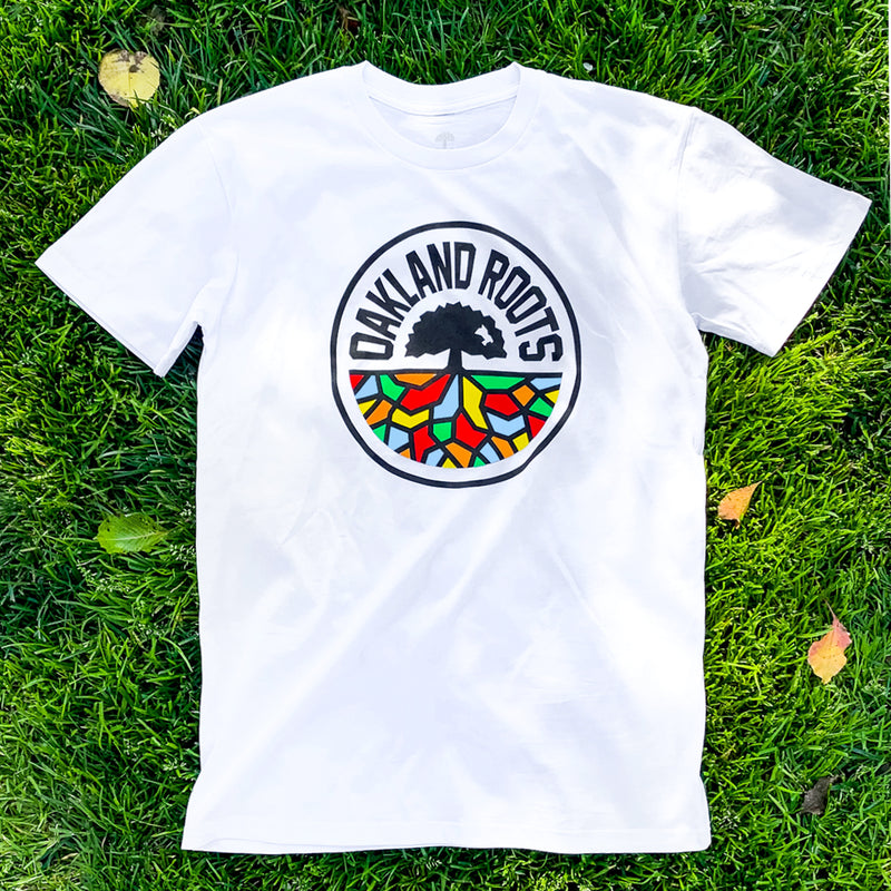 White t-shirt with full-color, round Oakland Roots logo on the chest lying on the grass.