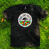 Black t-shirt with full-color, round Oakland Roots logo on the chest lying on the grass.
