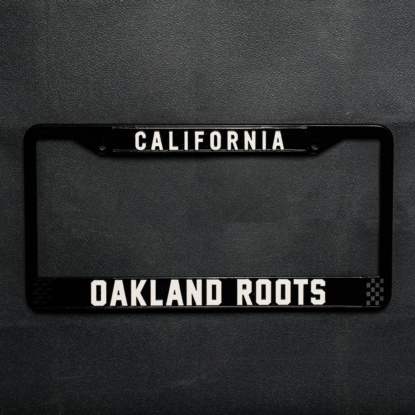 License plate holder with black rims with silver California wordmark on top and Oakland Roots logo on the bottom on a black vinyl background.