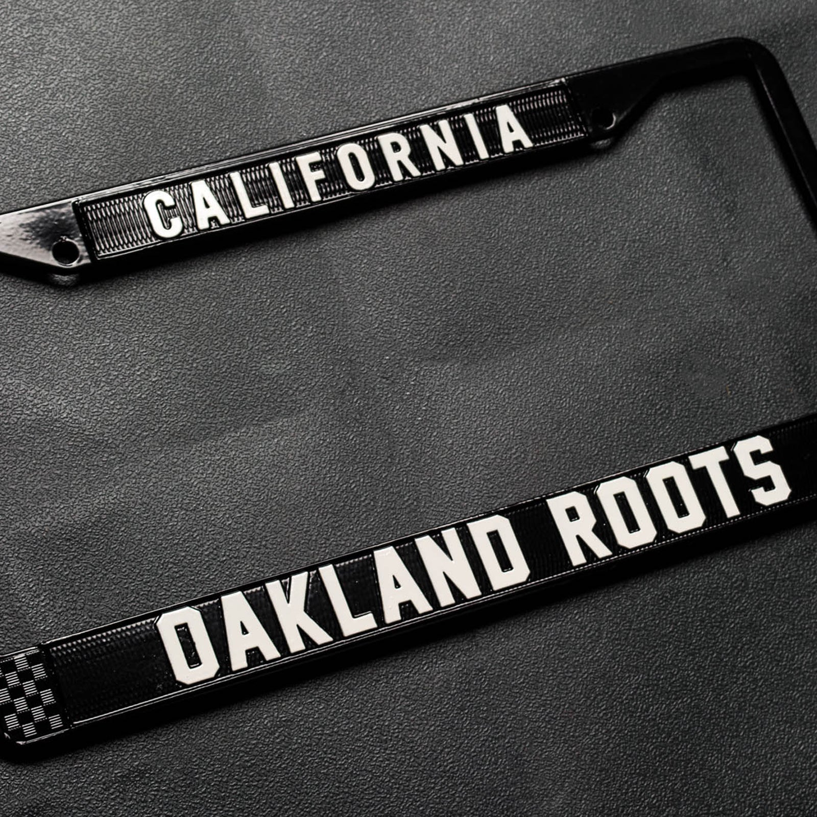 License plate holder with black rims with silver California wordmark on top and Oakland Roots logo on the bottom on a black vinyl background.