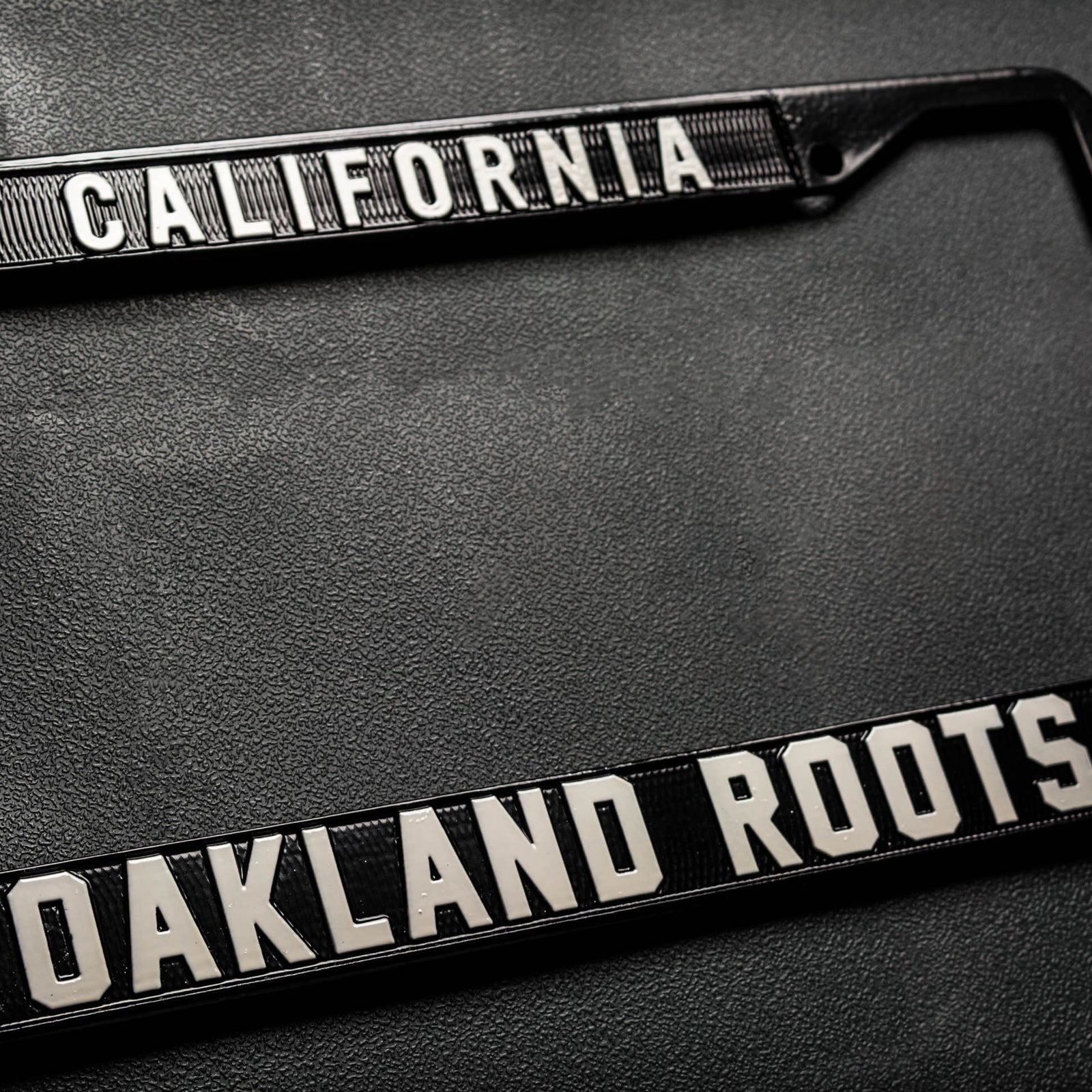 License plate holder with black rims with silver California wordmark on top and Oakland Roots logo on the bottom on a black vinyl background.