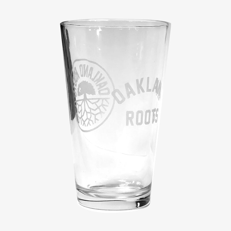 Beer pint glass with translucent white Oakland Roots logo crest on one side and OAKLAND ROOTS wordmark on the other.