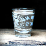 Liquor hot glass full of clear liquid with translucent Oakland Roots logo crest on shelf with faded background.