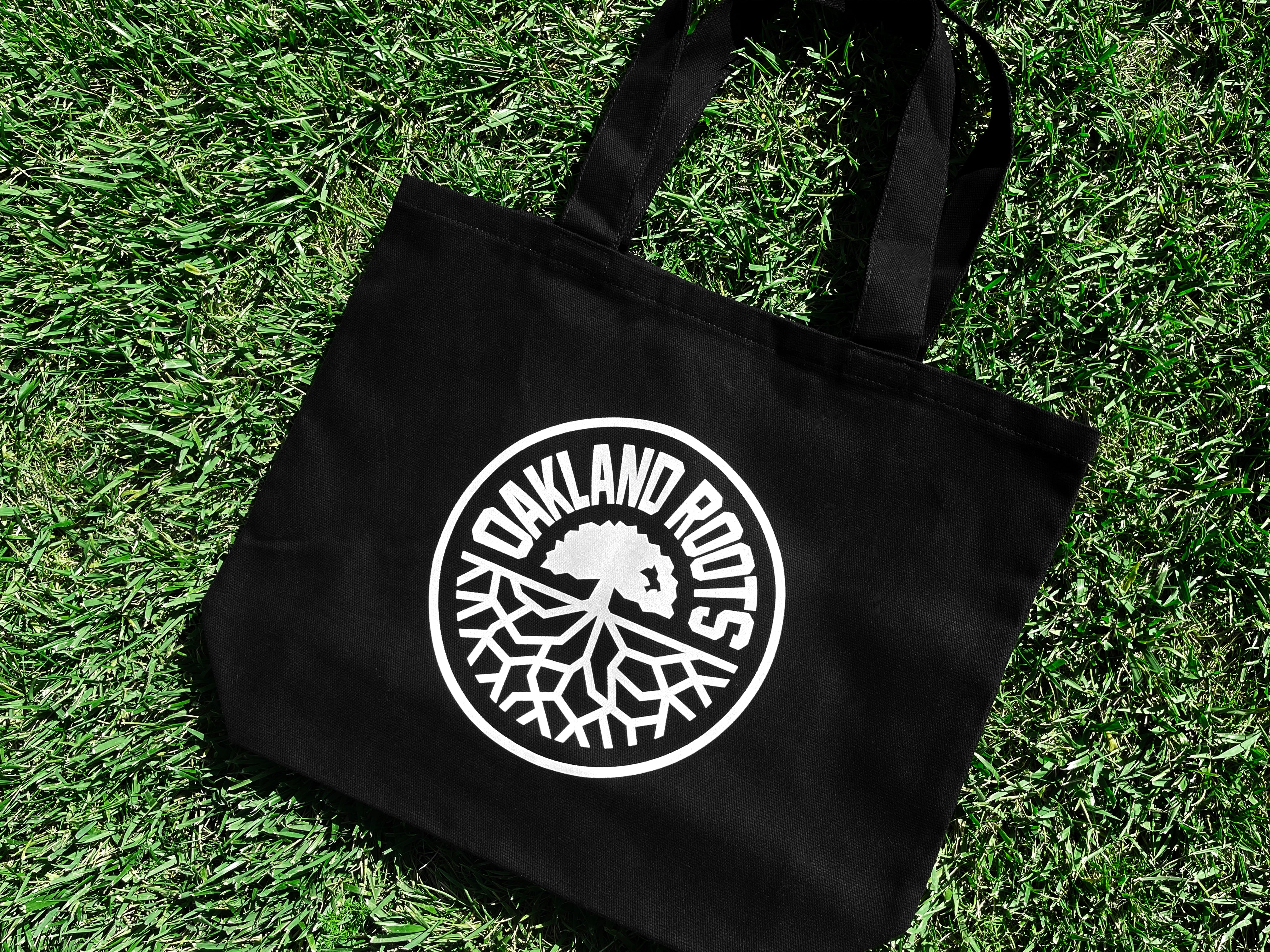 Close-up of black reusable shopping tote with large white Oakland Roots logo crest lying on grass.